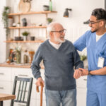 Smiling friendly in-home male nurse in uniform supporting an old man with a walking stick